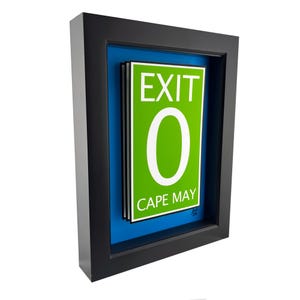 May include: Framed artwork featuring a green sign with the word "EXIT" above a large white "0" and the words "CAPE MAY" below. The sign is set against a blue background within a black frame.