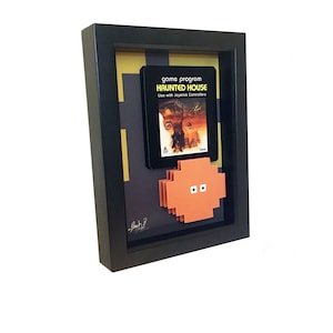 May include: Framed artwork featuring a vintage "Haunted House" video game cartridge. The black frame surrounds a pixelated orange character and a background with geometric shapes. The game cartridge is black with white and orange text.