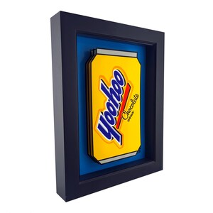 May include: A framed artwork featuring a yellow Yoohoo chocolate drink can. The can has blue and red text and is set against a blue background within a black frame. The artwork is a vibrant and eye-catching piece.