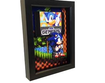 May include: A framed 3D artwork featuring a pixelated Sonic the Hedgehog character from the Sega Genesis video game. The artwork includes a Sega Genesis game cartridge with a picture of Sonic on the front.