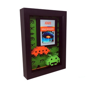 May include: A framed artwork featuring a vintage Atari Space Invaders game cartridge with a green background and two pixelated alien spaceships in 3D, one orange and one green.