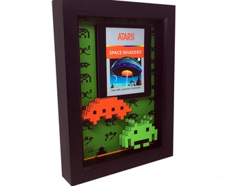 May include: A framed artwork featuring a vintage Atari Space Invaders game cartridge with a green background and two pixelated alien spaceships in 3D, one orange and one green.