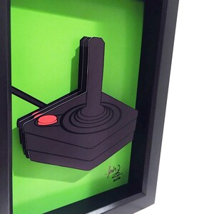 Puede incluir: Una ilustraci&oacute;n enmarcada de un joystick negro y rojo sobre un fondo verde. La firma del artista y el a&ntilde;o 2016 son visibles en la esquina inferior derecha.