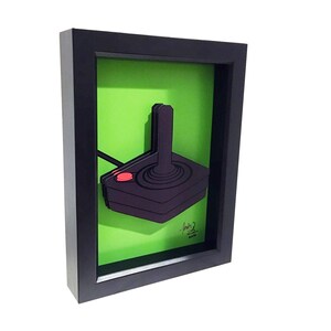 Puede incluir: Una obra de arte 3D enmarcada de un joystick negro y rojo sobre un fondo verde. La obra est&aacute; firmada "Josh 2 2019".