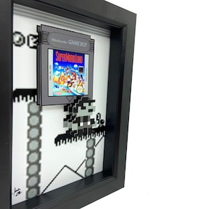 May include: A framed 3D pixel art piece featuring a Super Mario Land Game Boy cartridge and a pixelated Mario character. The frame is black and the background is white with a pixelated landscape.