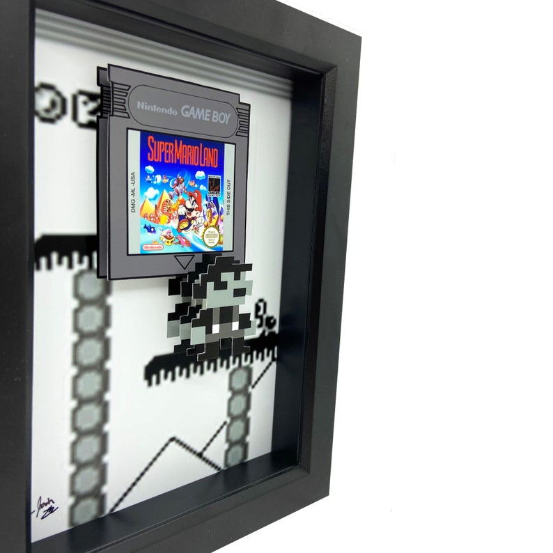 May include: A framed 3D pixel art piece featuring a Super Mario Land Game Boy cartridge and a pixelated Mario character. The frame is black and the background is white with a pixelated landscape.
