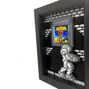 May include: A framed 3D pixel art piece featuring the classic Nintendo Game Boy game Metroid. The game cartridge is displayed in the frame, along with a pixelated depiction of Samus Aran, the protagonist of the game, walking across a pixelated background.