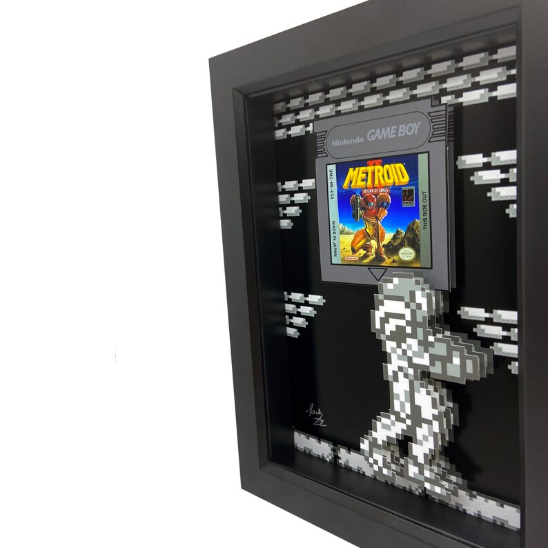 May include: A framed 3D pixel art piece featuring the classic Nintendo Game Boy game Metroid. The game cartridge is displayed in the frame, along with a pixelated depiction of Samus Aran, the protagonist of the game, walking across a pixelated background.