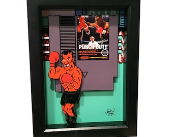 Puede incluir: Una pieza de arte pixelado en 3D enmarcada del clásico juego de Nintendo Punch-Out!! con el personaje principal, Little Mac, en una postura de boxeo. El marco es negro con un fondo verde y un cartucho de Nintendo morado.