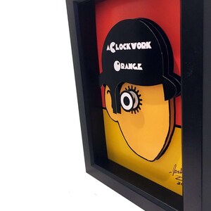 May include: A framed 3D pop art print of a character from the movie "A Clockwork Orange". The character is wearing a black bowler hat with the title of the movie printed on it. The character's face is yellow with a large black eye and a red background.