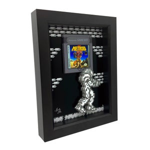 May include: A framed 3D pixel art piece featuring a character from the video game Metroid. The character is walking on a platform with a Game Boy cartridge for the game Metroid in the background. The frame is black and the art is in black and white.
