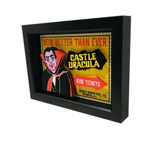 May include: A framed illustration of a vintage amusement park sign for a ride called "Castle Dracula". The sign is yellow with red and black lettering. A cartoon vampire with a red cape is featured on the sign.