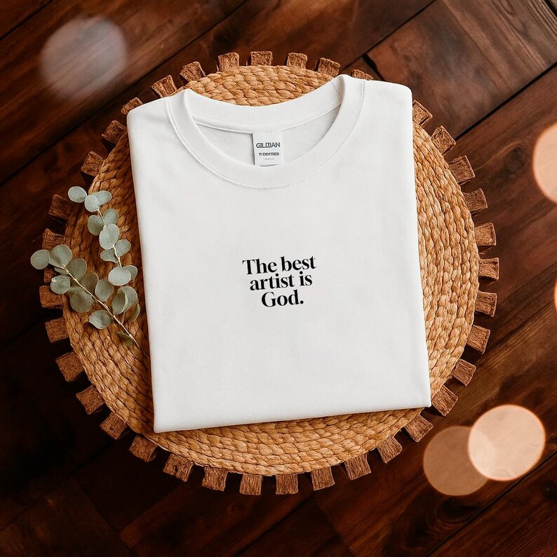 Pode incluir: Uma camiseta branca com o texto "The best artist is God". A camiseta &eacute; exibida em um sousplat circular tecido. A camiseta &eacute; da marca Gildan.