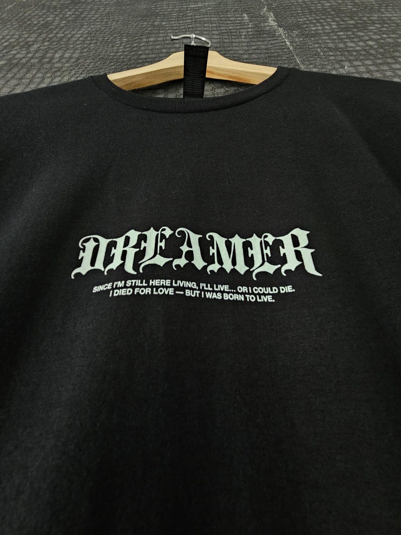 May include: Black crew neck sweatshirt with the word "DREAMER" in a gothic font. Below the word is the quote: "SINCE I'M STILL HERE LIVING, I'LL LIVE... OR I COULD DIE. I DIED FOR LOVE - BUT I WAS BORN TO LIVE."