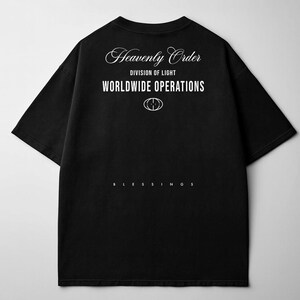 May include: Black t-shirt with white text that reads "Heavenly Order, Division of Light, Worldwide Operations" and a symbol. The word "Blessings" is printed at the bottom. The shirt has short sleeves and a crew neck.