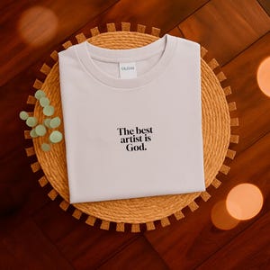 Pode incluir: Uma camiseta de cor clara com gola redonda com o texto "The best artist is God". A camisa &eacute; exibida em um sousplat circular tecido. O fundo &eacute; uma superf&iacute;cie de madeira escura.
