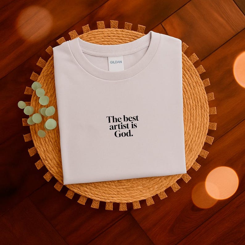 Pode incluir: Uma camiseta de cor clara com gola redonda com o texto "The best artist is God". A camisa &eacute; exibida em um sousplat circular tecido. O fundo &eacute; uma superf&iacute;cie de madeira escura.