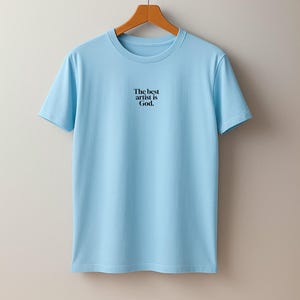 Pode incluir: T-shirt azul claro com gola redonda e o texto preto "The best artist is God.". A t-shirt est&aacute; pendurada num cabide de madeira sobre um fundo cinza claro.