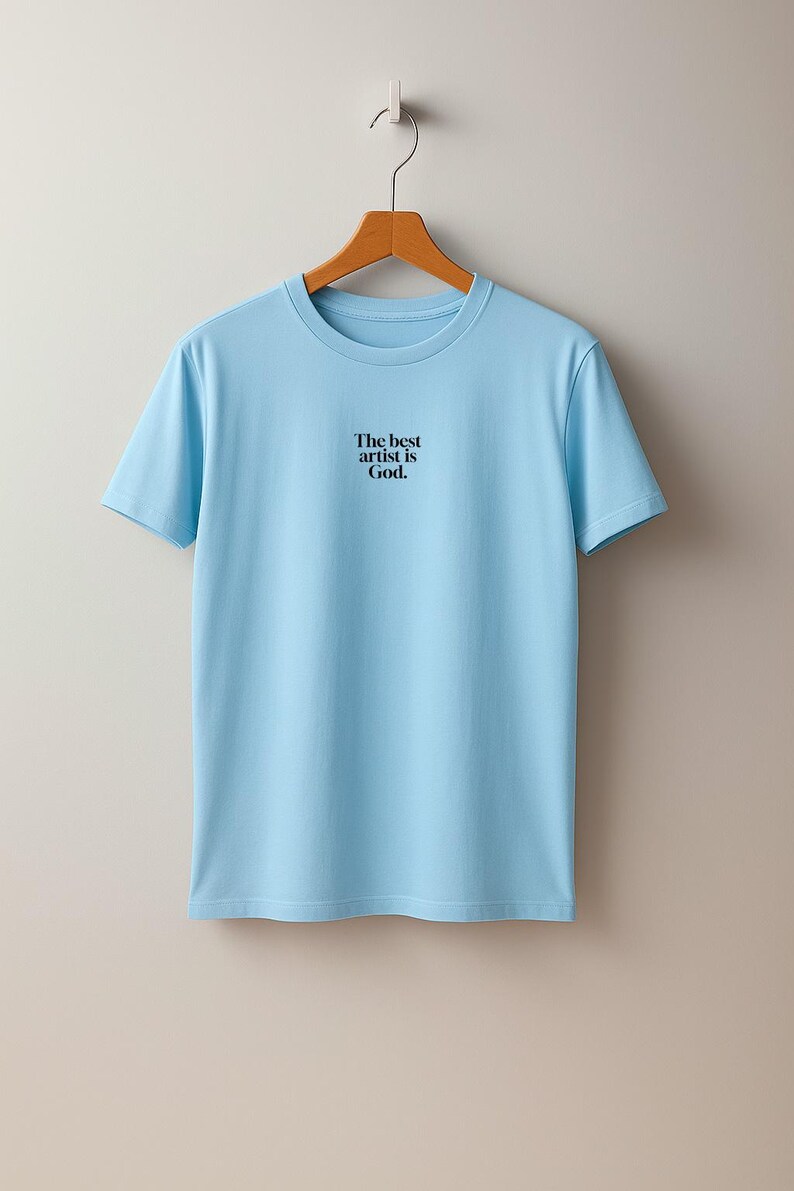 Pode incluir: T-shirt azul claro com gola redonda e o texto preto "The best artist is God.". A t-shirt est&aacute; pendurada num cabide de madeira sobre um fundo cinza claro.