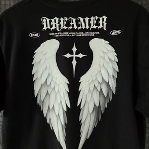 May include: Black t-shirt featuring a white graphic of angel wings and a cross. The word "DREAMER" is printed above the wings in a gothic font. Additional text includes "ESTD" and "2025", along with a quote about living and dying.