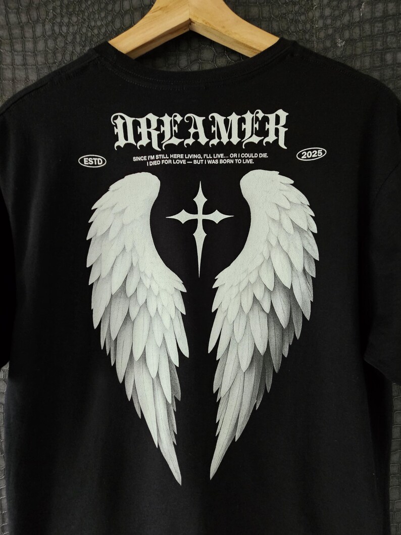 May include: Black t-shirt featuring a white graphic of angel wings and a cross. The word "DREAMER" is printed above the wings in a gothic font. Additional text includes "ESTD" and "2025", along with a quote about living and dying.