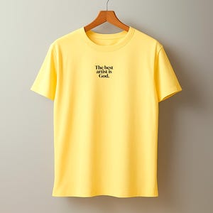 Pode incluir: Uma t-shirt amarelo brilhante com o texto preto "The best artist is God." exibida num cabide de madeira. A t-shirt tem gola redonda e mangas curtas.