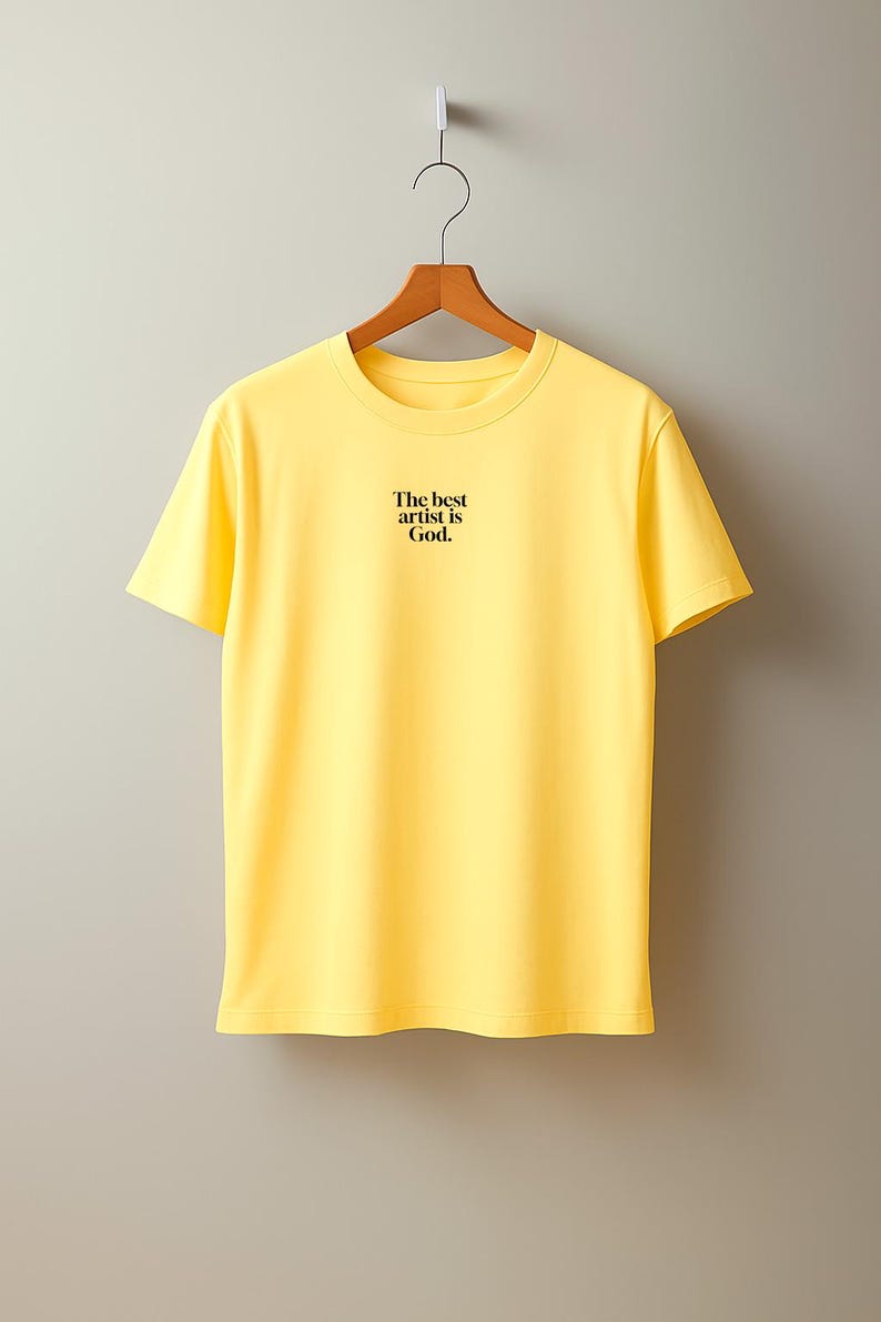 Pode incluir: Uma t-shirt amarelo brilhante com o texto preto "The best artist is God." exibida num cabide de madeira. A t-shirt tem gola redonda e mangas curtas.