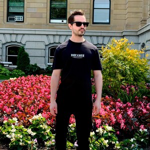 May include: A person wearing a black t-shirt with the word "DREAMER" in white script. The person is also wearing black sunglasses and black pants. The background features a building and a flower bed with pink and white flowers.