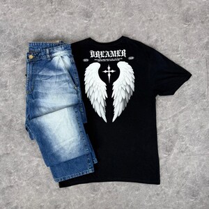 May include: A black t-shirt with white angel wings and a cross graphic, paired with blue denim jeans. The shirt features the word "DREAMER" at the top. The jeans are folded and have a button closure.