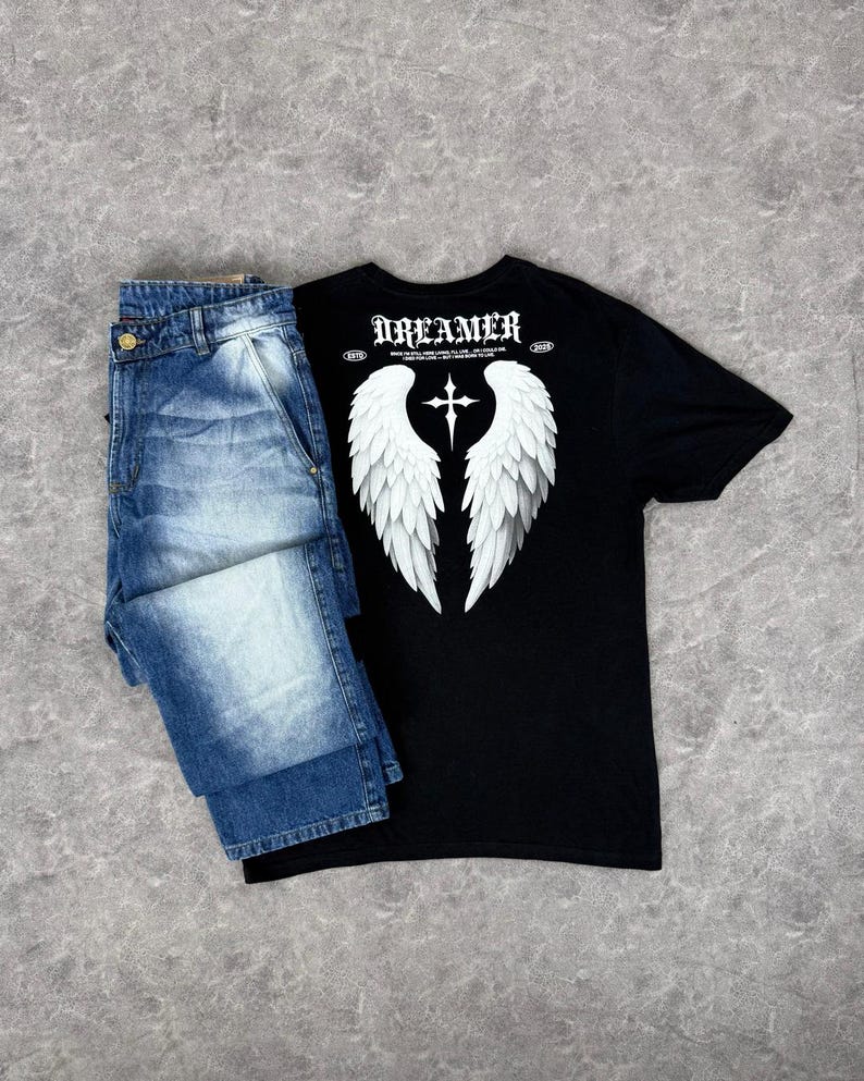 May include: A black t-shirt with white angel wings and a cross graphic, paired with blue denim jeans. The shirt features the word "DREAMER" at the top. The jeans are folded and have a button closure.
