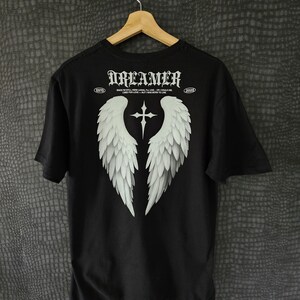 May include: Black t-shirt with a white graphic of angel wings and a cross. The word "DREAMER" is printed across the top in a gothic font. The shirt also has the text "ESTD 2023" and a quote.