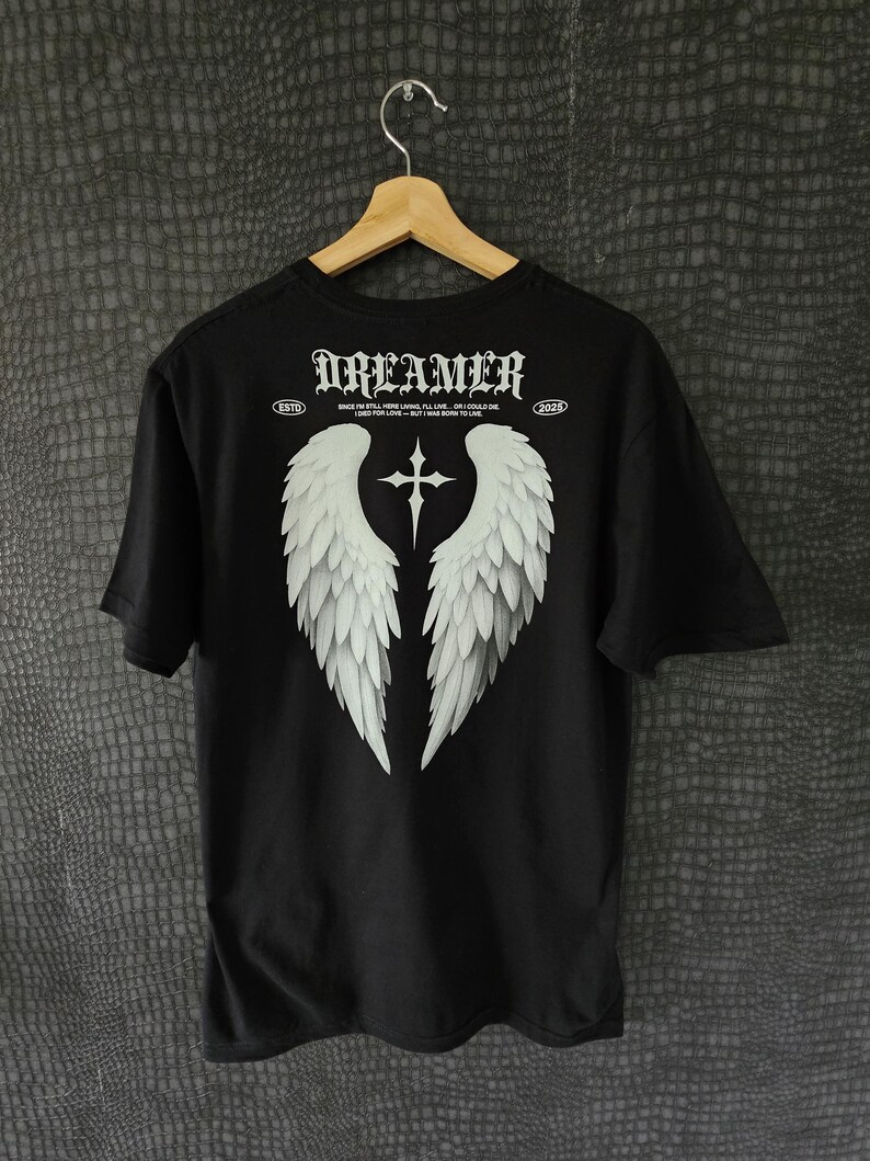 May include: Black t-shirt with a white graphic of angel wings and a cross. The word "DREAMER" is printed across the top in a gothic font. The shirt also has the text "ESTD 2023" and a quote.