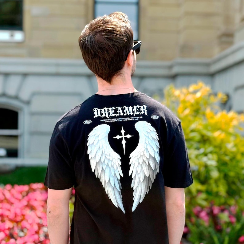 May include: Black t-shirt with the word "DREAMER" in white gothic lettering across the upper back. Below the text is a white cross and two large white angel wings. The shirt has a crew neck and short sleeves.