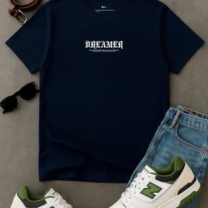 May include: Navy blue t-shirt with the word "DREAMER" in white gothic font. The shirt is paired with blue jeans, white and green sneakers, a brown belt, and black sunglasses. A casual, everyday outfit.