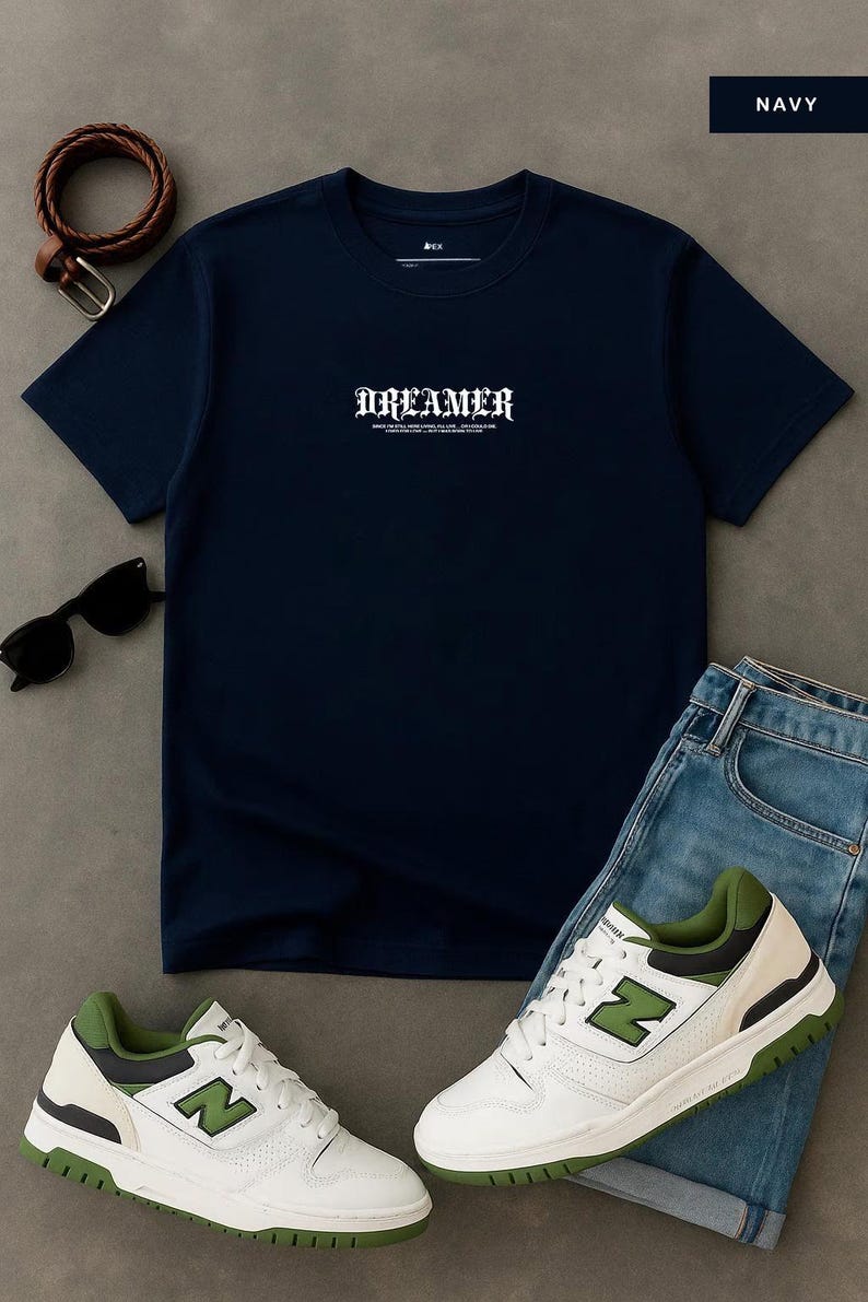 May include: Navy blue t-shirt with the word "DREAMER" in white gothic font. The shirt is paired with blue jeans, white and green sneakers, a brown belt, and black sunglasses. A casual, everyday outfit.