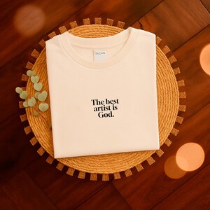 Pode incluir: Camiseta creme com o texto "The best artist is God". A camiseta &eacute; exibida em um sousplat redondo e tecido. O logotipo da Gildan &eacute; vis&iacute;vel na gola.