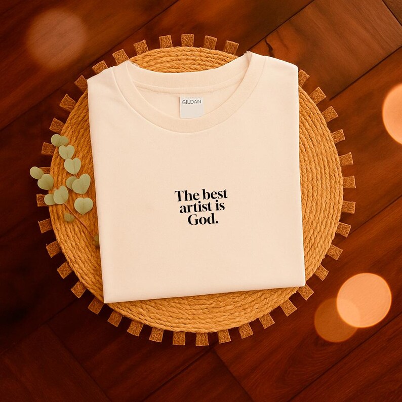 Pode incluir: Camiseta creme com o texto "The best artist is God". A camiseta &eacute; exibida em um sousplat redondo e tecido. O logotipo da Gildan &eacute; vis&iacute;vel na gola.