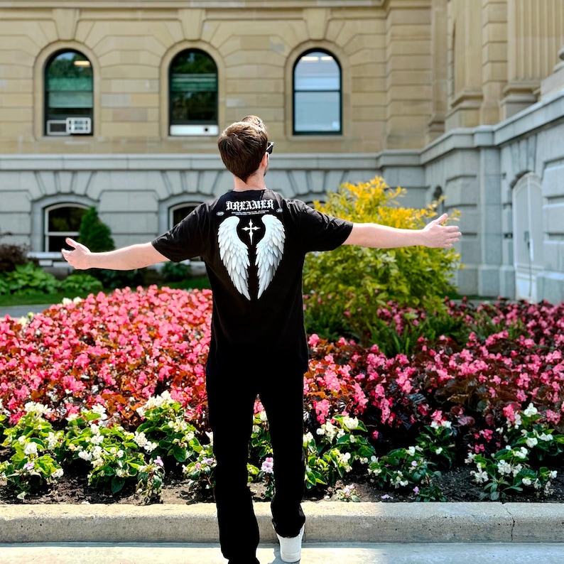 May include: Black t-shirt with the word "DREAMER" above a white wing and cross design. The person is wearing black pants and white shoes. The background includes a building and a flower bed.