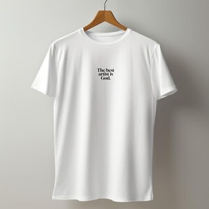 Pode incluir: Camiseta branca com o texto "The best artist is God" em preto, pendurada em um cabide de madeira. A camiseta &eacute; lisa e simples, com gola redonda e mangas curtas. O fundo &eacute; uma parede cinza claro.