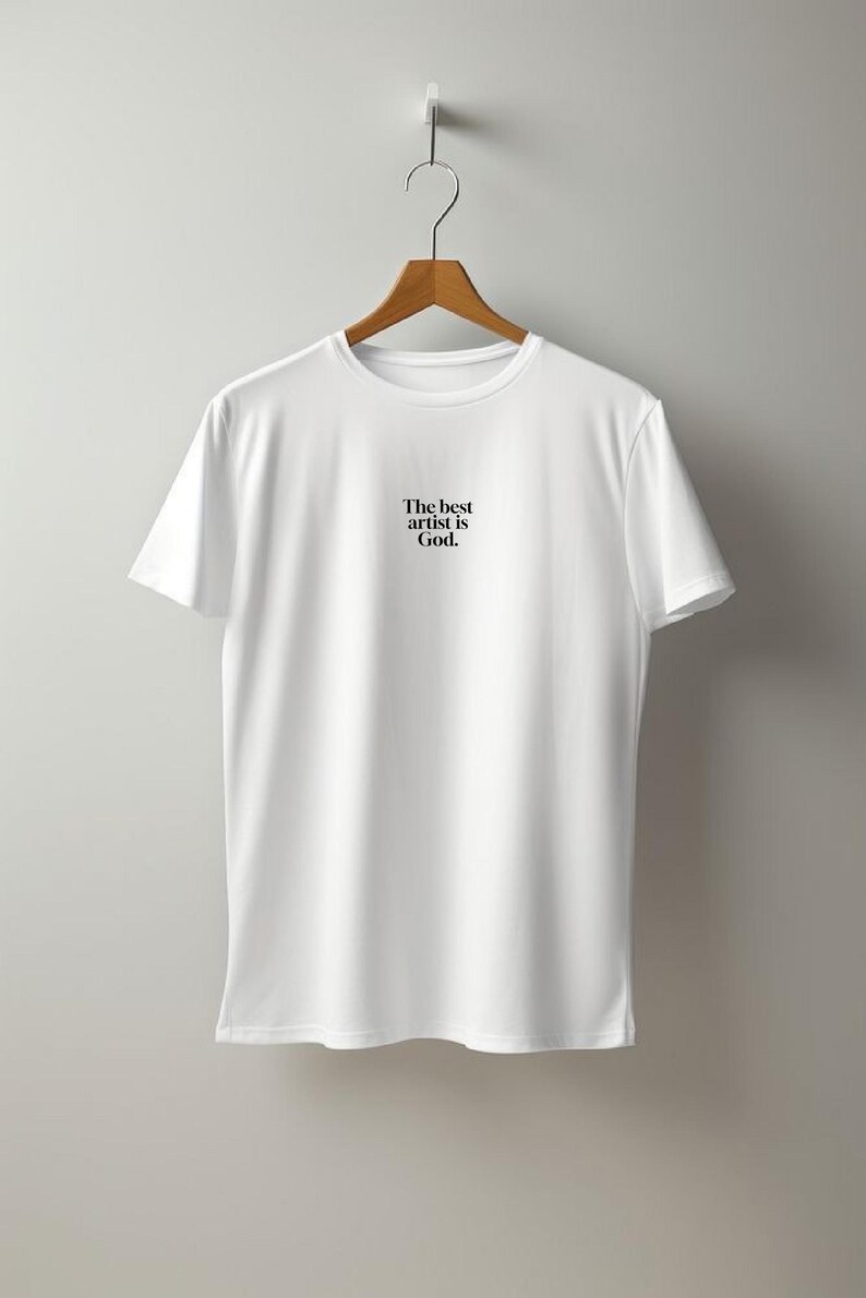 Pode incluir: Camiseta branca com o texto "The best artist is God" em preto, pendurada em um cabide de madeira. A camiseta &eacute; lisa e simples, com gola redonda e mangas curtas. O fundo &eacute; uma parede cinza claro.