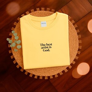 Pode incluir: Uma camiseta amarela dobrada com o texto "The best artist is God." A camiseta est&aacute; sobre um sousplat circular e tecido. A camiseta tem gola redonda e mangas curtas.