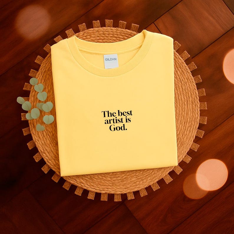Pode incluir: Uma camiseta amarela dobrada com o texto "The best artist is God." A camiseta est&aacute; sobre um sousplat circular e tecido. A camiseta tem gola redonda e mangas curtas.