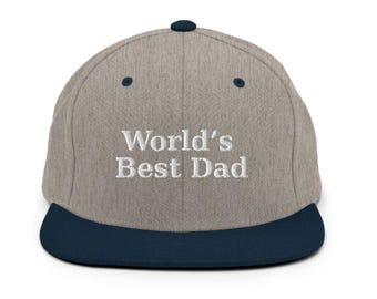 Op de afbeelding: Een baseballpet met een grijze kroon en een marineblauwe klep. De tekst "World's Best Dad" is in het wit op de voorkant geborduurd. De pet heeft een knoop bovenop en twee kleine marineblauwe accenten.