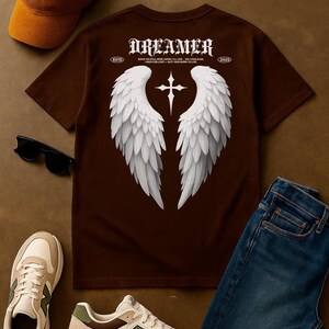 May include: Dark chocolate brown t-shirt with a white angel wing graphic and the word "Dreamer". The shirt is paired with a brown cap, sunglasses, sneakers, and blue jeans. The shirt has the text "ESTD 2025".