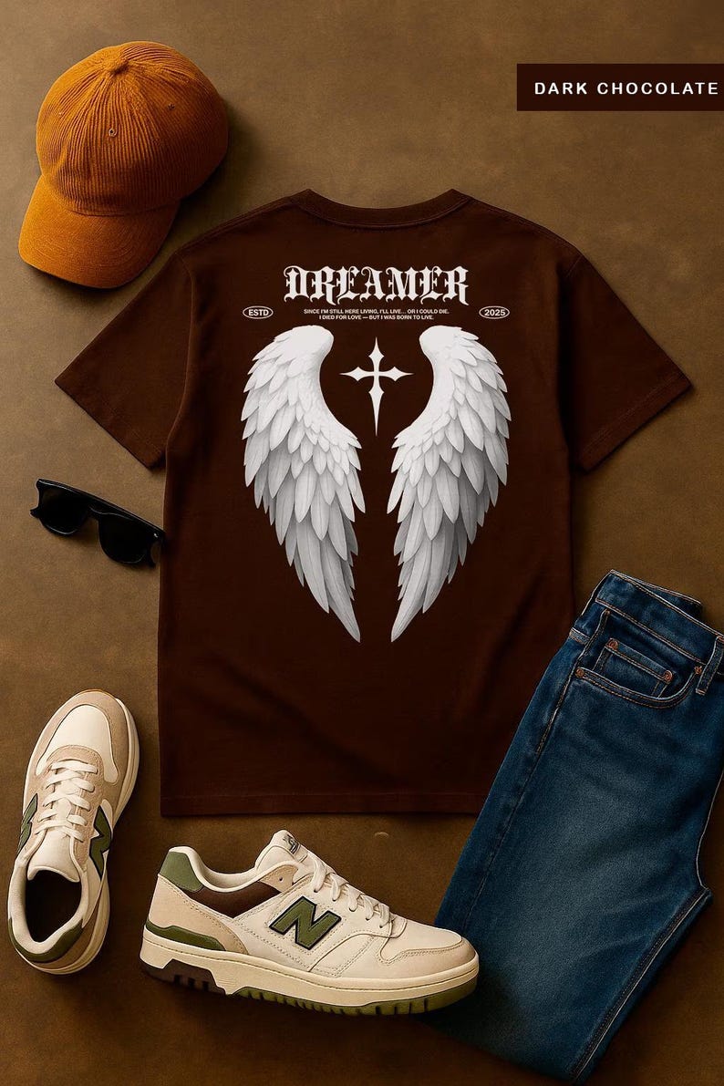 May include: Dark chocolate brown t-shirt with a white angel wing graphic and the word "Dreamer". The shirt is paired with a brown cap, sunglasses, sneakers, and blue jeans. The shirt has the text "ESTD 2025".