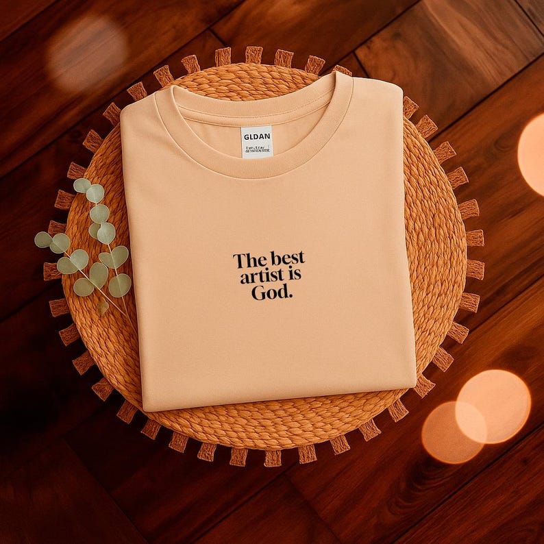 Pode incluir: Uma camiseta bege com o texto "The best artist is God" em preto. A camisa est&aacute; dobrada e colocada em um sousplat redondo e tecido. A camisa tem gola redonda e uma etiqueta "GILDAN".