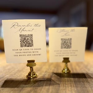 May include: Two cream-colored cards with gold-colored holders on a wooden surface. The cards have text and QR codes. One card reads "Remember this Moment..." and the other reads "Share the Love". The text on the cards encourages guests to share photos.