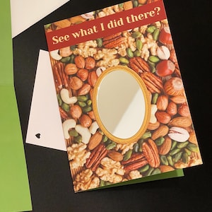 May include: Greeting card with a mirror and a background of mixed nuts. The card has the text "See what I did there?" in a red banner. A white envelope and a green card are also visible.