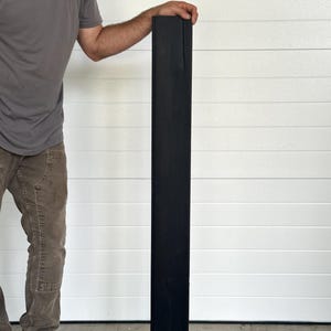 May include: A tall, rectangular, black wooden post. The post is held upright by a person. The wood grain is visible on the surface. The post is likely for construction or decorative purposes.