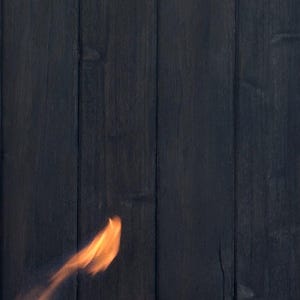 May include: A close-up shot of dark, vertical wooden planks. A blurred flame in shades of orange and yellow appears to the left, suggesting fire or heat. The wood grain is visible, adding texture.